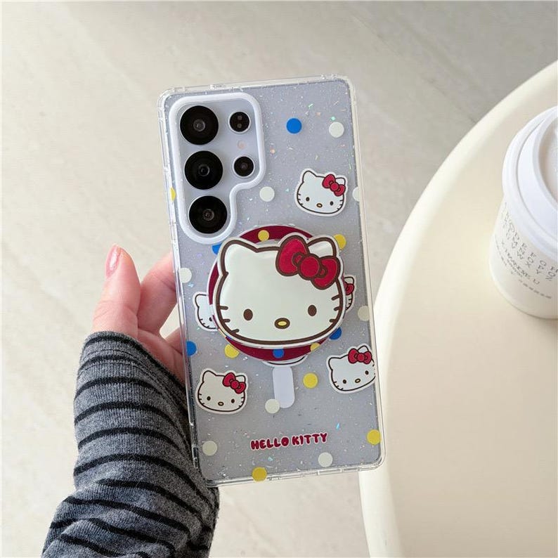 K&ouml;nnte beinhalten: Transparente Handyh&uuml;lle mit Hello Kitty-Design. Die H&uuml;lle zeigt ein gro&szlig;es Hello Kitty-Gesicht mit roter Schleife, umgeben von kleineren Hello Kitty-Gesichtern und Punkten. Die Worte "Hello Kitty" sind unten aufgedruckt.