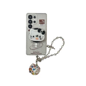 Puede incluir: Una funda transparente para tel&eacute;fono con un fondo brillante, con un dise&ntilde;o de Hello Kitty blanco con un lazo negro y un agarre emergente. La funda tiene un charm a juego con personajes de dibujos animados coloridos y una cadena de cuentas.