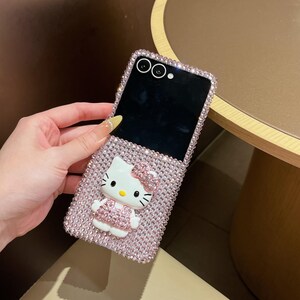 Puede incluir: Funda de tel&eacute;fono rosa con brillantes con una figura 3D de Hello Kitty. La funda est&aacute; cubierta de peque&ntilde;os brillantes rosas brillantes y tiene una pantalla negra. La figura de Hello Kitty tiene un lazo y un vestido rosa. La funda est&aacute; abierta.