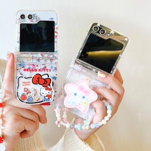 May include: Two clear phone cases, one open and one closed, featuring a Hello Kitty design. The open case displays a black screen and a Hello Kitty graphic with the text "Hello Kitty". The closed case has a star-shaped charm with a Hello Kitty face attached.