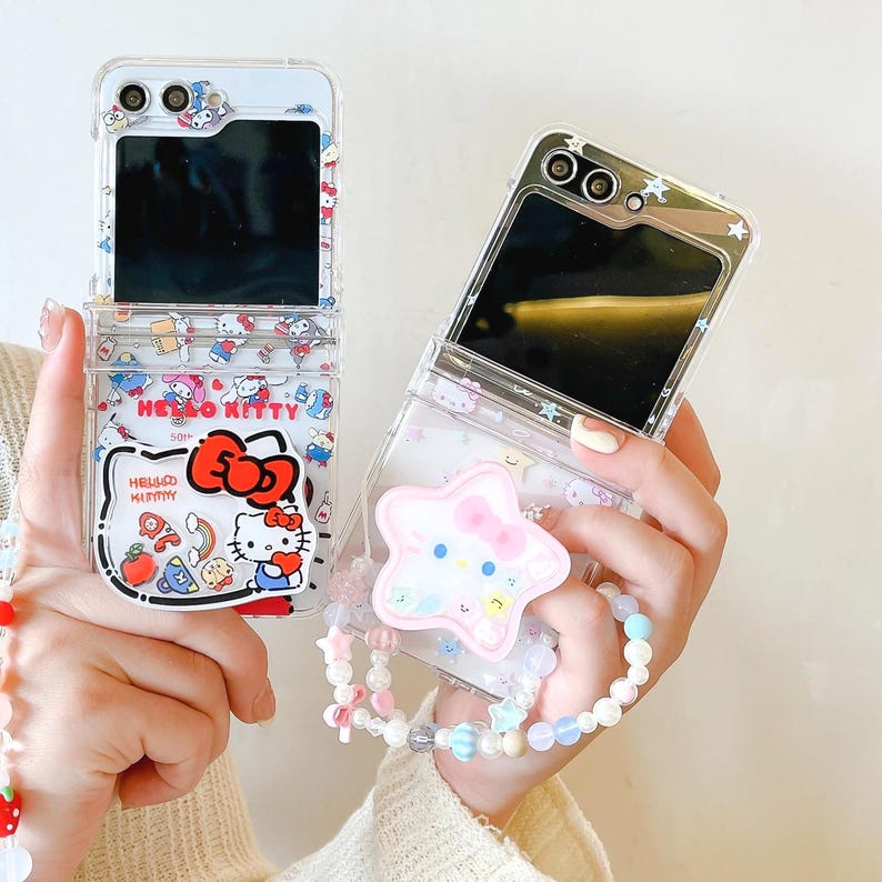 May include: Two clear phone cases, one open and one closed, featuring a Hello Kitty design. The open case displays a black screen and a Hello Kitty graphic with the text "Hello Kitty". The closed case has a star-shaped charm with a Hello Kitty face attached.