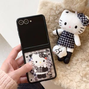 May include: Black flip phone case with a Hello Kitty design. The case features a clear, protective layer with a 3D Hello Kitty figure wearing a black and white houndstooth dress. A matching plush Hello Kitty doll with a bow and dress is also present.