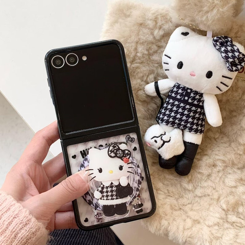 May include: Black flip phone case with a Hello Kitty design. The case features a clear, protective layer with a 3D Hello Kitty figure wearing a black and white houndstooth dress. A matching plush Hello Kitty doll with a bow and dress is also present.