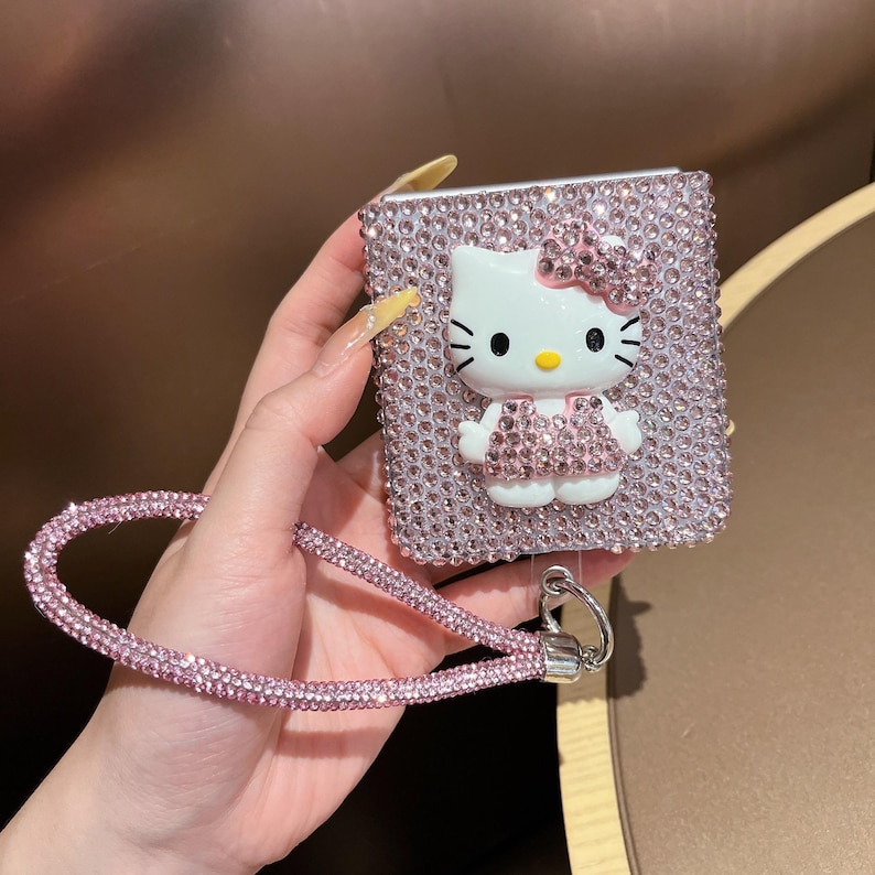 Puede incluir: Una funda rosa cubierta de pedrer&iacute;a con una figura 3D de Hello Kitty con un lazo y un vestido rosas. La funda tiene una correa de mu&ntilde;eca de pedrer&iacute;a rosa a juego sujeta con un cierre plateado. La funda es de forma cuadrada.