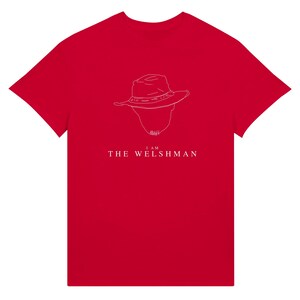 May include: A red t-shirt featuring a white outline of a hat and head, with the text "I AM THE WELSHMAN" below the illustration. The shirt is a solid, vibrant red color.