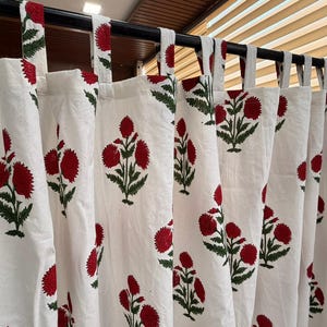 以下が含まれることがあります： 赤色の花柄と緑の葉の繰り返し模様が施された白いカーテン。カーテンは黒いロッドから吊り下げられており、上部に布製のループが付いています。花柄はブロックプリントスタイルです。