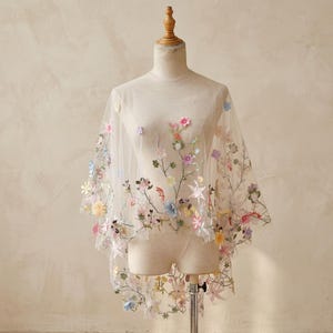May include: A sheer, off-white tulle capelet featuring delicate, multi-colored floral embroidery. The garment displays a variety of small, three-dimensional fabric flowers in shades of pink, blue, yellow, and green, scattered across the lightweight fabric.