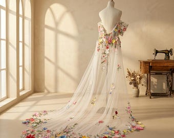 May include: A sheer, floor-length gown on a mannequin, featuring an off-the-shoulder neckline and a long, flowing train. The garment is decorated with colorful, three-dimensional floral appliqués in shades of red, blue, yellow, and pink.