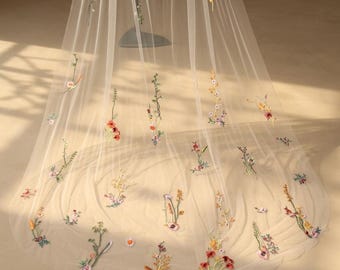 May include: A sheer, ivory-colored veil with delicate floral embroidery. The veil features a variety of colorful embroidered flowers, including red, pink, yellow, and purple, scattered across the fabric. The veil is likely for a wedding or special occasion.