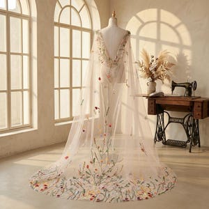 May include: A floor-length, sheer beige cape displayed on a mannequin, featuring a deep V-neckline and intricate, colorful floral embroidery scattered across the fabric, with a dense cluster of wildflowers detailed along the sweeping hemline.