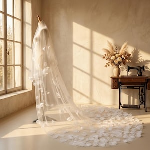 Pode incluir: Um longo véu de noiva branco e transparente drapeado sobre um manequim, apresentando pétalas de tecido branco espalhadas por todo o comprimento do tule, estendendo-se pelo chão numa cauda, criando uma aparência suave, em camadas e fluida.