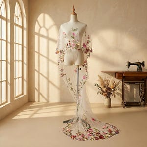 Puede incluir: Una capa de tul blanco transparente con intrincados bordados florales en hilo rosa, rojo y verde. La prenda cubre un maniquí y se extiende en una larga cola fluida adornada con patrones botánicos dispersos y flores de colores variados.