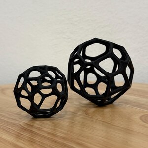 Könnte beinhalten: Zwei schwarze, hohle, kugelförmige Objekte mit einer komplexen, miteinander verbundenen geometrischen Gitterstruktur. Die Kugeln weisen ein sich wiederholendes Muster aus fünfeckigen und sechseckigen Öffnungen auf, was ein leichtes, offenes Rahmendesign mit matter Oberfläche ergibt.
