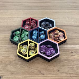 Könnte beinhalten: Ein Set aus sieben ineinandergreifenden sechseckigen Behältern mit schwarzem Innenraum und farbigen Rändern in Rot, Weiß, Orange, Pink, Gelb, Olivgrün und Dunkelblau. Jeder Behälter ist in zwei Abschnitte unterteilt und mit kleinen, nummerierten Spielmarken gefüllt.