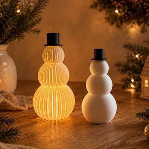 Könnte beinhalten: Zwei Schneemann-Figuren mit schwarzen Zylindern. Der größere Schneemann links hat einen gerippten, durchscheinenden weißen Körper, der von innen warm beleuchtet wird. Der kleinere Schneemann rechts hat eine glatte, einfarbig weiße, matte Oberfläche.