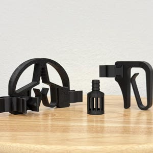 Könnte beinhalten: Ein Set aus drei schwarzen Kunststoffkomponenten, bestehend aus einer halbkreisförmigen Halterung mit mehrzinkiger Basis, einem kleinen zylindrischen Filter mit gerippter Düse und einer hakenförmigen Klemme mit rechteckigem Befestigungspunkt.