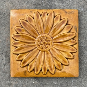 May include: A ceramic tile with a raised, embossed design of a yellow daisy flower. The tile has a rustic, glazed finish.