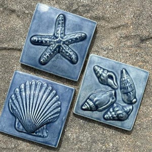 May include: Three blue ceramic tiles with raised designs of seashells and a starfish. The tiles are square and have a glossy finish.