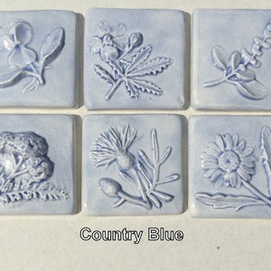 May include: Six blue ceramic tiles with embossed floral designs. Each tile features a different flower or plant, including a daisy, a thistle, a fern, and a wildflower. The tiles are labeled "Country Blue".