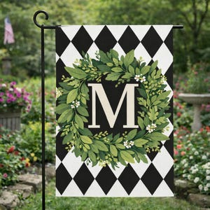 May include: A garden flag featuring a black and white diamond pattern background. A green wreath with white flowers surrounds a large white letter "M". The flag is hanging on a black metal stand in a garden setting.