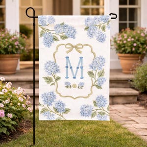 May include: A white garden flag featuring blue hydrangea flowers and green leaves. The flag has a decorative frame with a bow and the letter "M" in blue. The flag is displayed on a black metal stand in a garden setting.