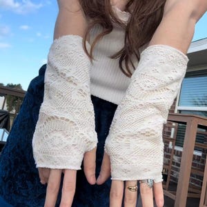 May include: Off-white lace fingerless gloves. The gloves are made of a delicate, open-weave lace fabric. The gloves cover the hand up to the knuckles and extend up the forearm. The background shows a blue sky.