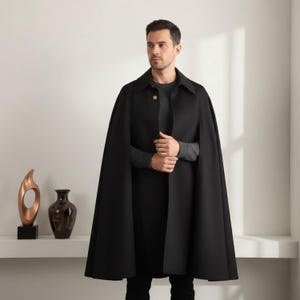 May include: A black cape with a collared neckline and a single gold button closure. The cape is made of a smooth, flowing fabric and has a classic design. The person is wearing a gray sweater underneath.