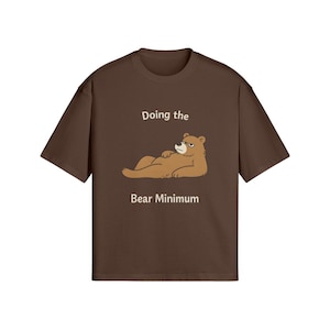 Puede incluir: Camiseta marrón con la frase "Doing the Bear Minimum" y una ilustración de un oso de dibujos animados. El oso está tumbado con una expresión de mal humor. La camiseta es de color marrón liso.