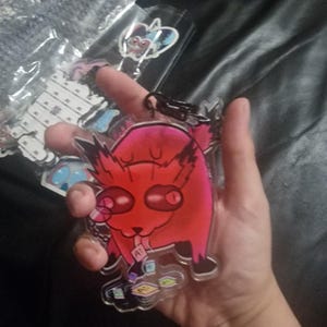 May include: A clear acrylic keychain featuring a vibrant red and pink creature with large dark eyes and spiky ears. The character is depicted standing over several small, colorful square tiles that each display the letter A. A black metal clasp is attached.