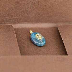 May include: A blue and gold pendant with an oval-shaped, blue stone and a smaller, oval-shaped, gold-colored stone in the center. The pendant has a gold bail. The pendant is displayed in a brown box.