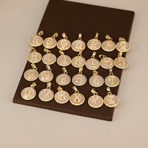 May include: A collection of gold-colored initial pendants, each featuring a letter from the alphabet. The pendants have a circular design with a white or light pink background and are displayed on a brown surface.