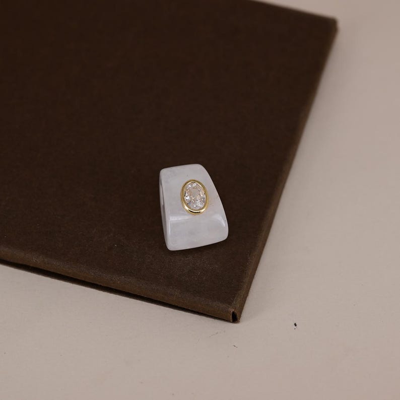 Pu&ograve; includere: Un anello bianco, traslucido e di forma trapezoidale con una gemma trasparente dal taglio ovale al centro, incastonata in una lunetta color oro lucido. L'anello presenta una finitura liscia e lucida e una fascia spessa che si restringe verso l'alto.