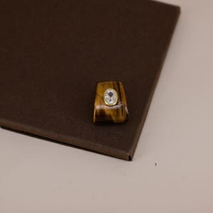 Pu&ograve; includere: Un anello caratterizzato da una pietra occhio di tigre color marrone dorato lucida con una superficie liscia e curva. Al centro della pietra si trova una piccola gemma trasparente di forma ovale incastonata in un castone d'oro giallo brillante.