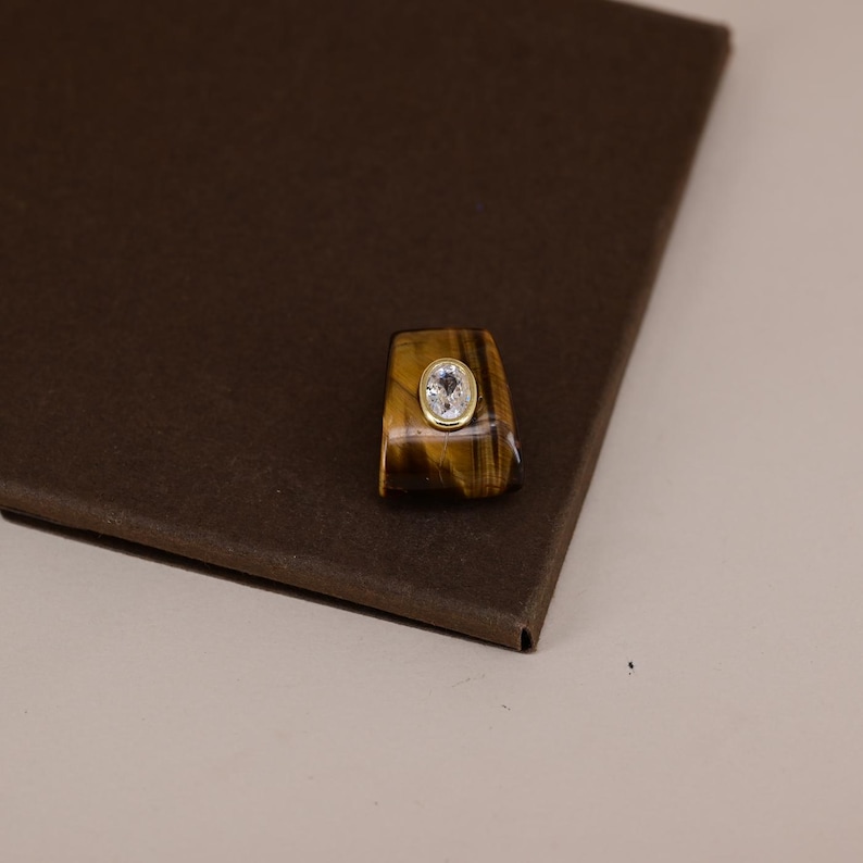 Pu&ograve; includere: Un anello caratterizzato da una pietra occhio di tigre color marrone dorato lucida con una superficie liscia e curva. Al centro della pietra si trova una piccola gemma trasparente di forma ovale incastonata in un castone d'oro giallo brillante.