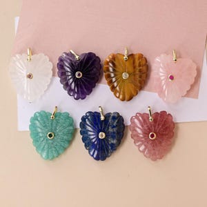 May include: Seven heart-shaped pendants carved from various gemstones, including white quartz, amethyst, tiger's eye, rose quartz, green aventurine, lapis lazuli, and strawberry quartz. Each pendant features a gold-tone bail and a central gemstone.