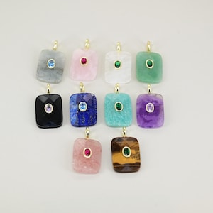 May include: A collection of rectangular gemstone pendants in various colors, including gray, pink, white, green, black, blue, turquoise, purple, and brown. Each pendant features a smaller, contrasting gemstone set in a gold-colored bezel.