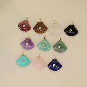 Peut inclure: Une collection de pendentifs en forme de coquillage de différentes couleurs : rouge, marron, blanc, noir, violet, sarcelle, vert, turquoise, rose et bleu marine. Chaque pendentif a une bélière dorée et une petite pierre ronde et transparente au centre.