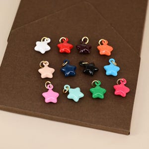 Puede incluir: Una colección de colgantes en forma de estrella de colores, incluyendo blanco, rojo, morado, naranja, melocotón, azul marino, negro, azul claro, rosa, turquesa, verde y rosa intenso. Cada colgante tiene un lazo dorado.