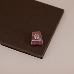 Pu&ograve; includere: Una pietra rosa di forma trapezoidale con una superficie liscia e lucida, caratterizzata da una gemma trasparente di forma ovale incastonata in una cornice di metallo dorato. La pietra rosa presenta sottili inclusioni bianche e una qualit&agrave; traslucida, mentre la gemma chiara &egrave; sfaccettata.