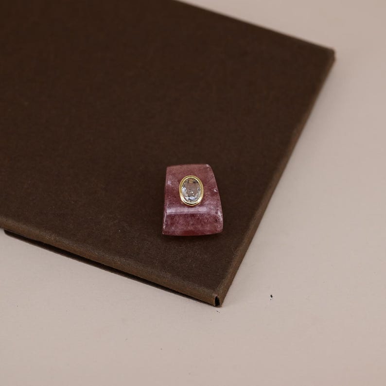 Pu&ograve; includere: Una pietra rosa di forma trapezoidale con una superficie liscia e lucida, caratterizzata da una gemma trasparente di forma ovale incastonata in una cornice di metallo dorato. La pietra rosa presenta sottili inclusioni bianche e una qualit&agrave; traslucida, mentre la gemma chiara &egrave; sfaccettata.