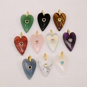 May include: Ten heart-shaped pendants made of various polished gemstones, each featuring a gold-toned bail with small clear stones and a central gold-toned heart inlay. The stones include green, black, brown, red, pink, white, purple, blue, and marbled white.