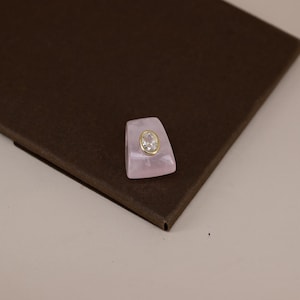 Pu&ograve; includere: Un anello caratterizzato da una fascia trapezoidale in materiale traslucido rosa pallido. Al centro della parte anteriore, una gemma trasparente dal taglio ovale &egrave; incastonata in una lunetta in metallo dorato, creando un contrasto con il rosa.