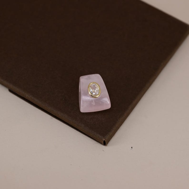 Pu&ograve; includere: Un anello caratterizzato da una fascia trapezoidale in materiale traslucido rosa pallido. Al centro della parte anteriore, una gemma trasparente dal taglio ovale &egrave; incastonata in una lunetta in metallo dorato, creando un contrasto con il rosa.