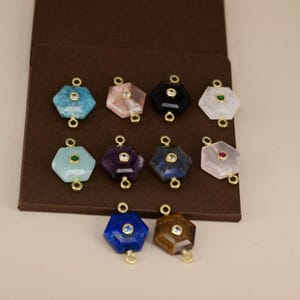 Op de afbeelding: Een verzameling zeshoekige edelsteenhangers in verschillende kleuren, waaronder blauw, roze, zwart en wit. Elke hanger heeft een goudkleurig metalen frame en een kleine edelsteen in het midden. De hangers worden gepresenteerd op een bruin oppervlak.