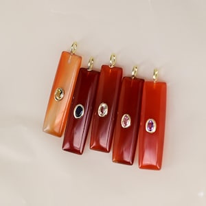 May include: Five rectangular pendants in shades of orange and red, each with a gold-colored loop at the top. Each pendant features a small, oval-shaped gemstone in various colors, including green, blue, and pink.