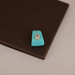 Pu&ograve; includere: Una pietra turchese trapezoidale con una gemma trasparente di forma ovale al centro, incastonata in una lunetta color oro. Il turchese presenta venature naturali visibili di colore marrone chiaro che contrastano con la superficie blu brillante.