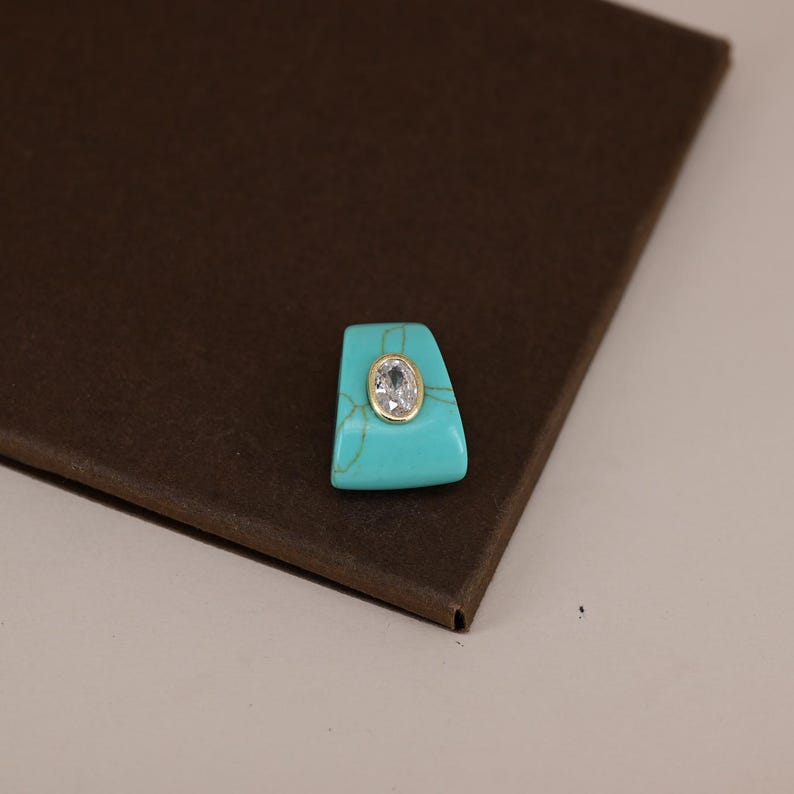 Pu&ograve; includere: Una pietra turchese trapezoidale con una gemma trasparente di forma ovale al centro, incastonata in una lunetta color oro. Il turchese presenta venature naturali visibili di colore marrone chiaro che contrastano con la superficie blu brillante.