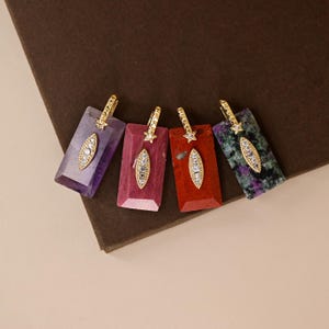 May include: Four rectangular pendants featuring polished gemstones in purple, pink, red, and a speckled green and purple mix. Each pendant has a gold-toned bail with a small star accent and a central marquise-shaped gold inlay set with clear stones.