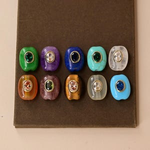 Könnte beinhalten: Zehn ovale Perlen in zwei Reihen angeordnet, jede mit einem zentralen, goldgerahmten Edelstein. Die Perlen zeigen verschiedene Farben wie Grün, Lila, Blau, Türkis, Transparent, Orange, Braun und Weiß mit unterschiedlichen Texturen und Oberflächen.
