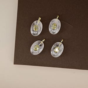 May include: Four oval-shaped pendants with a gold bail and a central yellow gemstone. The pendants are set in a clear, slightly cloudy material, possibly quartz or resin. The background is a dark brown.